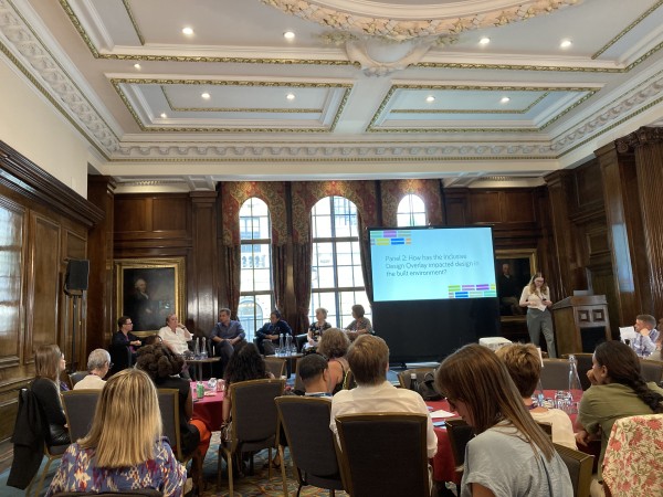 A photo taken from behind a group of people sitting down and listening to someone talk on stage