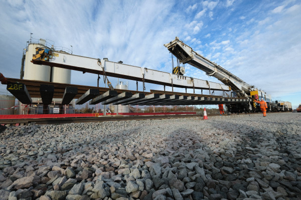 A large rail-mounted crane lowers a long section of railway track components over newly laid rails. Workers in high-visibility orange safety clothing guide the suspended load, while gravel ballast fills the foreground. Industrial storage tanks and fencing are visible in the background beneath a dramatic blue sky with streaked clouds.