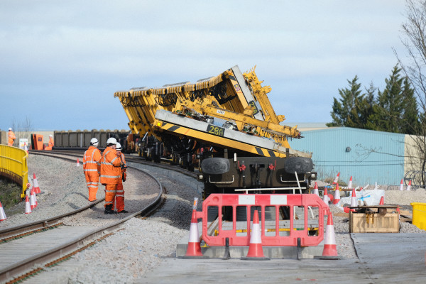 Rail workers in high-visibility orange safety clothing stand beside a curved railway track as a large yellow track-laying machine operates on newly installed rails. Traffic cones and safety barriers mark the construction area, with industrial buildings and trees visible in the background under a cloudy sky.